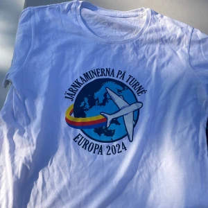 Vit t-shirt Järnkaminerna Europa 2024 Djurgården Sofialäktaren  - Vit t-shirt med tryck på bröstet: Järnkaminerna på turné Europa 2024. Motivet visar ett flygplan som flyger över en blå jordglob med röd och gul slinga. Rund hals och korta ärmar, perfekt för dig som gillar supporterkläder. Passar perfekt att ha på Sofialäktaren 
