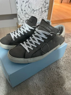 Philippe Model grå sneakers i skinn - Snygga sneakers från Philippe Model i grått skinn med vita detaljer och vita snören. Klassisk låg modell med rund tå och vit sula. Märkets logga på plösen och sidan. Perfekta för dig som gillar stilrena och exklusiva sneakers. Box kommer med.