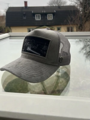 Grå truckerkeps med mesh och tryck - Snygg grå truckerkeps med böjd skärm och mesh på sidorna och bak för extra ventilation. Framtill sitter ett svartvitt tryck med stadsmotiv. Justerbar rem baktill för perfekt passform. Perfekt för dig som vill ha en stilren och urban look.