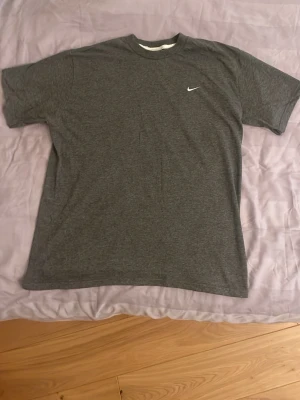 Grå Nike t-shirt med logga - Snygg grå t-shirt från Nike med klassisk vit broderad swoosh på bröstet. T-shirten har rund hals och korta ärmar, perfekt för en avslappnad och sportig stil. Tillverkad i mjuk bomull för skön komfort. Kan gå ner i pris vid snabb försäljning.