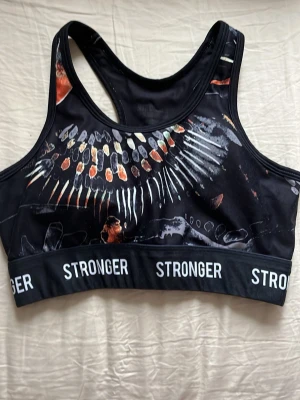 Mönstrad sport-bh från Stronger - Snygg sport-bh från Stronger i svart med färgglatt abstrakt mönster i orange, vitt och grått. Resårband med logga runt om och brottarrygg för extra stöd. Perfekt för träning och aktiva dagar. Kan inte hitta storleken men skulle gissa på S. Säljer då den inte passar mig