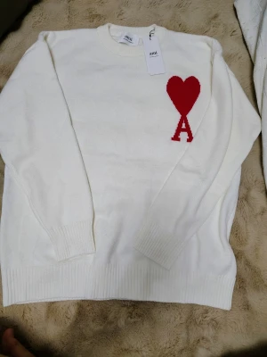 Vit stickad tröja från Ami Paris - Vit stickad tröja från Ami Paris med röd hjärtlogga och bokstaven A på bröstet. Tröjan har rund halsringning, långa ärmar och ribbade muddar vid ärmslut och nederkant. Perfekt för en stilren och trendig look.