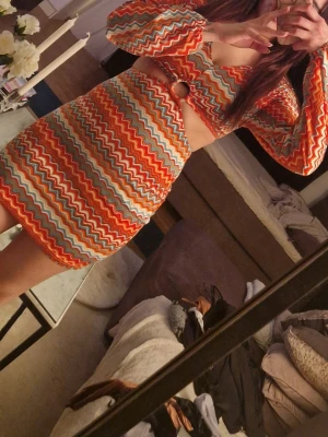 Färgglad klänning med zickzackmönster - Trendig klänning med lång ärm och färgglatt zickzackmönster i orange, rött, blått och vitt. Toppen har en rund ringdetalj framtill och är tillverkad i stickat material. Perfekt för dig som gillar färg och retrovibbar.