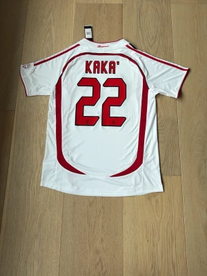 AC Milan Kaka' 22 bortatröja Adidas - Säljer en vit AC Milan bortatröja från Adidas med Kaka' och nummer 22 på ryggen. Tröjan har röda och svarta detaljer, Champions League-märke och Bwin som sponsor på bröstet. Perfekt för dig som älskar fotboll och vill ha en ikonisk supportertröja.