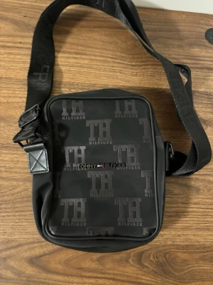 Svart axelväska Tommy Hilfiger - Snygg svart axelväska från Tommy Hilfiger med justerbar rem och präglad logotyp över hela väskan. Väskan har dragkedja och är perfekt för att bära det viktigaste. Klassisk och stilren design med TH-mönster.