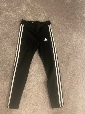 Svarta Adidas leggings med vita ränder. Barnstorkek 11-12y!! - Snygga svarta leggings från Adidas med klassiska vita ränder längs benen och Adidas-logga på låret. Tight passform och stretchigt material som passar perfekt till träning eller chill. Ikonisk sportig look som alltid funkar.