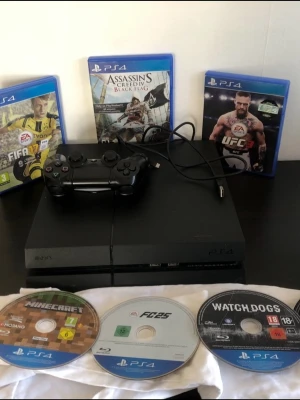 Playstation 4 med handkontroll och spel - Svart Playstation 4 med en svart DualShock 4-handkontroll och laddkabel. Flera PS4-spel ingår, bland annat FIFA, Assassin's Creed Black Flag, UFC 3, Minecraft, FC25 och Watch Dogs. 