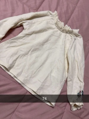 Vit blus med volangkrage och broderi - Söt vit blus med volangkrage och långa ärmar. Blusen har diskret struktur i tyget och ett litet broderi med blommor på ena ärmen. Perfekt för en trendig och romantisk look.