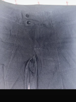 Svarta utsvängda kostymbyxor med dubbla knappar - Säljer ett par svarta kostymbyxor med hög midja och två snygga knappar framtill. Byxorna har utsvängda ben och är tillverkade i ett mjukt material som känns bekvämt. Perfekta för dig som gillar stilrena och klassiska plagg.
