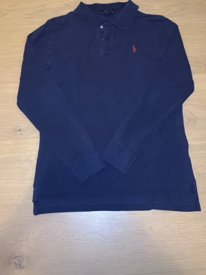 Marinblå långärmad piké från Polo Ralph Lauren - Klassisk mörkblå långärmad pikétröja från Polo Ralph Lauren med röd broderad logga på bröstet. Tröjan har krage, knappar vid halsen och ribbade muddar vid ärmsluten. Perfekt för en stilren och avslappnad look. Skicket är mycket bra men har en jätte liten defekt (se sista bilden)!