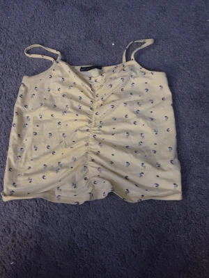 Gul blommig topp från XOXO Friends - Söt gul topp med tunna axelband från XOXO Friends. Toppen har små blommiga mönster i lila och svart samt rynkade detaljer framtill. Perfekt för varma dagar och enkel att matcha med jeans eller kjol.