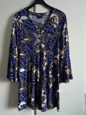 Blåvit mönstrad klänning/tunika med snörning - Klänning/tunika med blått och vitt paisleymönster, v-ringning med snörningsdetalj framtill och långa utsvängda ärmar. Endast använd några gånger. Inga fel på den.