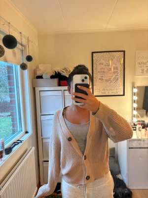 Beige ribbad kofta med knappar - Säljer en beige ribbad kofta från lager med stora mörka knappar framtill. Koftan har v-ringning, långa ärmar och breda muddar vid ärmslut och nederkant. Perfekt för lager på lager och enkel att matcha med olika outfits.