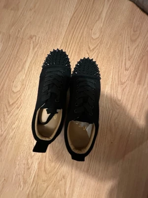 Svarta sneakers med nitar från Louboutin - Svarta sneakers från Christian Louboutin med coola svarta nitar på tån. Skorna har snörning och är gjorda i mocka med beige insida. Perfekta för dig som vill sticka ut med en edgy och lyxig stil. Jag köpte de på en second hand butik i italien.