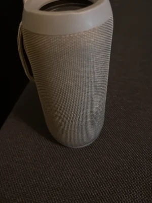 Grå cylinderformad bärbar högtalare - Snygg bärbar högtalare i grått tygmaterial med cylinderformad design. Högtalaren har en praktisk bärrem på sidan och robust plast i topp och botten. Perfekt för att ta med sig överallt och spela musik trådlöst.