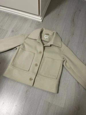 Beige kappa från Kappahl  - Stilren beige kappa från Kappahl med stora fyrkantiga fickor framtill och klassisk krage. Kappan har tre knappar och är i ett mjukt ull-liknande material. Köpt på kappahl. Lite nopprig vid ärmarna. 