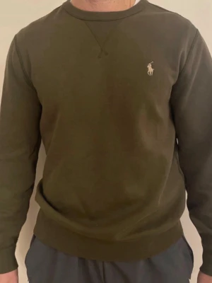 Olivgrön sweatshirt från Ralph Lauren - Snygg olivgrön sweatshirt från Ralph Lauren med klassisk rund halsringning och broderad logga på bröstet. Tröjan har ribbade muddar vid ärmslut och nederkant. Perfekt för en avslappnad och stilren look.