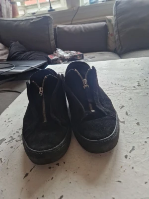 Svarta axel arigatos - Säljer ett par svarta sneakers i mocka med coola dragkedjor framtill istället för klassisk snörning. Skorna har låg profil och platt sula, vilket ger en clean och stilren look. Perfekta till jeans eller chinos för en avslappnad vibe.dock lite sönder men priset är inte satt så det går att förhandla