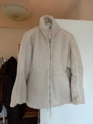 Beige teddyjacka från H&M - Mysig beige teddyjacka från H&M med hög krage och hel dragkedja framtill. Jackan har två stora fickor och är fodrad för extra värme. Perfekt för kyliga dagar och ger en avslappnad, trendig look.
