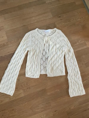 Vit stickad kofta med rosett - Söt vit stickad kofta med hålmönster och knytband i halsen. Koftan har långa, lätt utsvängda ärmar och kortare passform. Perfekt att slänga över en topp för en romantisk look.