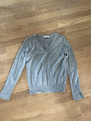Grå v-ringad stickad tröja från Zara - Snygg och enkel grå stickad tröja från Zara med v-ringning och långa ärmar. Tröjan har en smal passform och ribbade muddar vid ärmslut och nederkant. Perfekt att matcha med jeans eller kjol för en clean look.