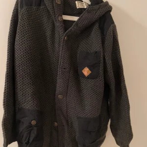 Grågrön kofta med huva för herr, storlek XL, Originals - Mysig grågrön kofta med huva och knappar framtill. Från Originals. Koftan har tre fickor med knappstängning och en liten patch på bröstfickan. Grovstickad struktur och svarta detaljer på axlar och fickor ger en cool kontrast.  Bra skick se defekter på bild.