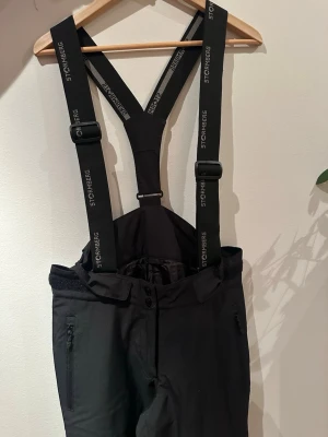 Svarta skidbyxor - Svarta skidbyxor från Stormberg med justerbara hängslen och dragkedjor vid fickorna. Byxorna har hög midja och är perfekta för vinterns alla äventyr. Materialet är tåligt och håller dig varm.