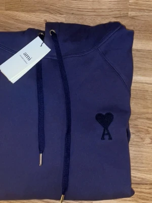 Mörkblå hoodie från Ami Paris - Snygg mörkblå hoodie från Ami Paris med klassisk huva och dragsko. På bröstet finns Ami Paris ikoniska hjärtemblem i matchande färg. Hoodien har känguruficka framtill och ribbade muddar vid ärmar och nederkant. Perfekt för en avslappnad och trendig look. 