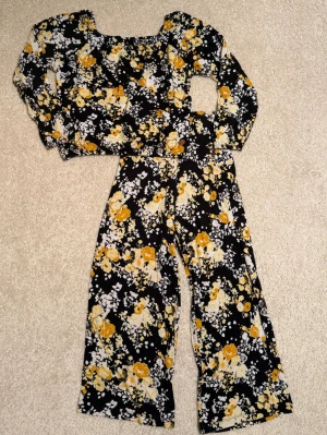 Blommigt loungewear-set i svart och gult, retro. - Snyggt loungewear-set med långärmad topp och vida byxor i svart tyg med gult och vitt blommönster. Setet har en avslappnad passform och mjukt material som passar perfekt för chill dagar hemma. Toppen har rundad hals och byxorna har resår i midjan.