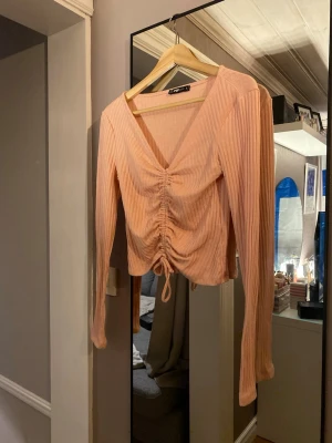 Ribbad rosa omlottopp från Sister - Trendig ribbad topp i ljusrosa från Sister med omlottdesign och snörning framtill. Långärmad modell med v-ringning och croppad passform. Perfekt för dig som gillar detaljer och en feminin look.