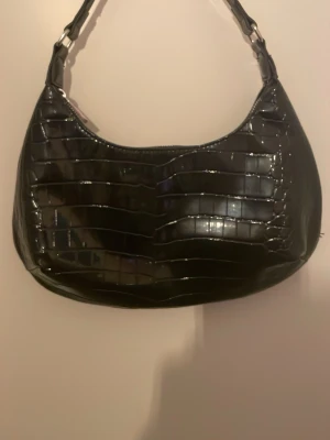 Svart croco handväska i syntet - Trendig svart handväska med croco-mönster och glansig finish. Väskan har en rundad form och ett kort axelband, perfekt för att lyfta din outfit. Gjord i syntetmaterial med stilrena detaljer.