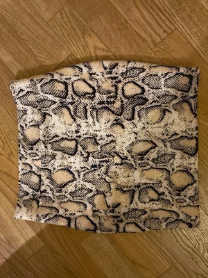 Beige kortkjol med ormmönster - Säljer en beige och svart kortkjol med trendigt ormmönster. Kjolen har en rak passform och är tillverkad i ett mjukt syntetmaterial som sitter snyggt på kroppen. Perfekt för dig som vill sticka ut lite extra.