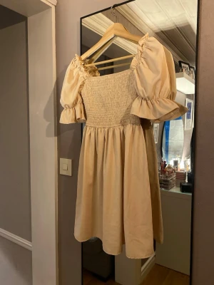 Beige kort klänning med puffärmar - Supersöt beige kort klänning med puffiga kortärmar och smockad överdel. Klänningen har en luftig kjol och volangdetaljer vid ärmsluten. Perfekt för sommarens alla tillfällen och ger en romantisk vibe.