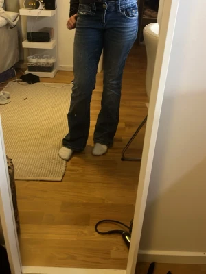 Blå Gina tricot bootcut jeans - Säljer ett par blå jeans från True Religion med ikonisk broderad ficka bak och snygg bootcut passform. Jeansen har klassisk femficksdesign, normal midja och är tillverkade i slitstarkt denim. Perfekta för dig som vill ha en tidlös och trendig look. Knapen är borta på på ena fickan