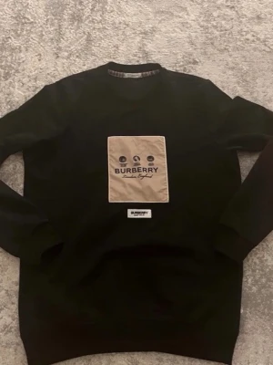Svart sweatshirt från Burberry - Stilren svart sweatshirt från Burberry med beige patch på bröstet och tryckt logga samt texten 'London, England'. Rund halsringning och ribbade muddar. Perfekt för dig som gillar exklusiva streetwear-plagg.