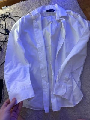 Vit klassisk skjorta från H&M - Säljer en vit klassisk skjorta från H&M med lång ärm och knappar framtill. Skjortan har en tidlös design med krage och är gjord i bomull för en skön känsla. Perfekt att styla lager på lager eller bära som den är.