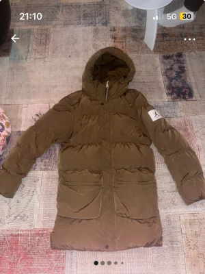 Brun dunjacka från Jordan med huva - Säljer en brun dunjacka från Jordan med huva och två stora fickor framtill. Jackan har en patch med Jumpman-loggan på ärmen och är perfekt för kalla dagar. Klassisk pufferstil med lång ärm och dragkedja framtill. Nypris: 2899kr