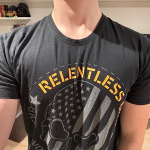 Svart t-shirt GASP - Svart t-shirt med tryck i gult och grått från GASP. Framsidan har ett stort motiv med texten 'RELENTLESS' och en döskalle med amerikansk flagga. Baksidan har vertikal gul text. Rund hals och korta ärmar. Skön bomullskänsla, perfekt till gymmet. Nypris 349kr. Mitt pris 450kr. Kolla även in mina andra annonser!