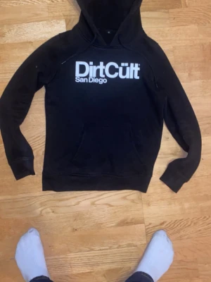 Svart Dirt Cult hoodie med tryck - Svart hoodie från Dirt Cult San Diego med stor vit logga på bröstet. Klassisk huva och känguruficka framtill. Tillverkad i mjuk bomullsmix, perfekt för en avslappnad streetwear-look.