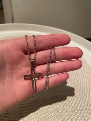 Kedjehalsband med kors i silverfärg - Snyggt halsband med en kedja i silverfärg och ett stort korsformat hänge. Hänget är ihåligt och har en modern, stilren design. Perfekt accessoar för dig som gillar minimalistiska smycken och vill ha något som sticker ut. Kedjan är 50 cm.