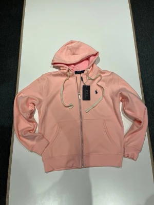 Ljusrosa hoodie från Polo Ralph Lauren - Snygg ljusrosa hoodie från Polo Ralph Lauren med dragkedja framtill, två fickor och vit dragsko i huvan. Klassisk broderad logga på bröstet och mjukt material som känns skönt mot huden. Perfekt för en avslappnad och trendig look.