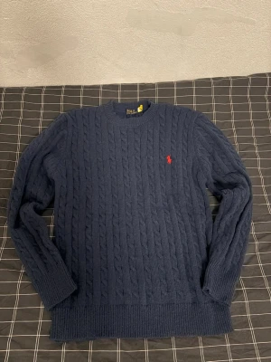 Mörkblå stickad tröja Polo Ralph Lauren - Klassisk mörkblå kabelstickad tröja från Polo Ralph Lauren med rund hals och röd broderad logga på bröstet. Tröjan har långa ärmar och ribbade muddar vid ärmslut och nederkant. Perfekt för dig som gillar stilren och tidlös design. Använd 2 gånger. Är i nyskick, hör av dig om du har några frågor.Kan gå ner till 500 vid snabb affär behöver  få såldt snabbt