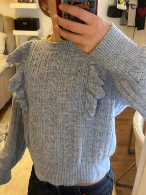 Ljusblå stickad tröja med volanger - Supersöt ljusblå stickad tröja från Lindex med dekorativa volanger längs axlarna och ett fint hålmönster framtill. Tröjan har rund halsringning, ribbade muddar och är perfekt för dig som gillar en gullig och trendig stil.