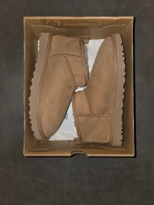 Bruna UGG boots med fårskinn - Klassiska bruna UGG boots i mocka med mjukt vitt fårskinnsfoder. Skorna har rund tå, platt sula och UGG-logga bak. Perfekta för kalla dagar och superbekväma att ha på sig.