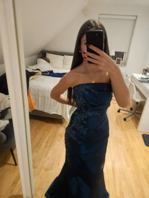 Marinblå axelbandslös långklänning - Snygg marinblå långklänning med axelbandslös design och glittriga detaljer. Klänningen har figurnära passform och elegant fall. Perfekt för bal eller festliga tillfällen.