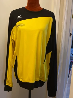 Gul och svart sporttröja från Erima - Sportig långärmad tröja från Erima i gult och svart med tryckt logga på bröstet. Tröjan har rund halsringning och är tillverkad i ett lätt och funktionellt material, perfekt för träning eller match.
