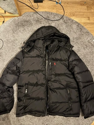 Svart pufferjacka från Polo Ralph Lauren - Snygg svart pufferjacka från Polo Ralph Lauren med huva och dragkedja framtill. Jackan har två dragkedjeförsedda fickor och det klassiska röda Polo-logot på bröstet. Perfekt för kalla dagar och riktigt stilren look.
