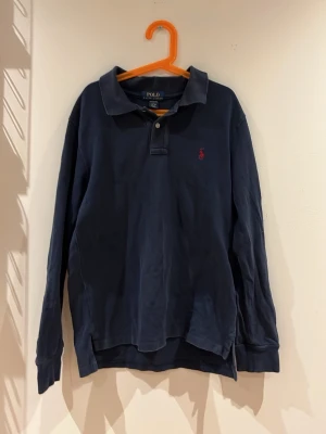 Mörkblå långärmad piké från Polo Ralph Lauren - Klassisk mörkblå långärmad pikétröja från Polo Ralph Lauren med krage, knappar vid halsen och den ikoniska röda logotypen broderad på bröstet. Perfekt för en stilren och avslappnad look.