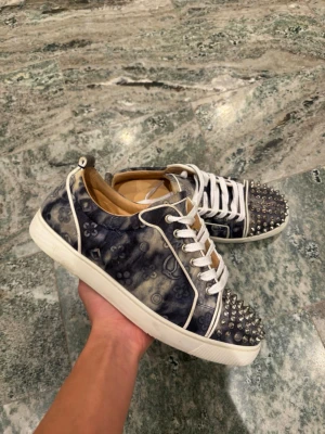 Louboutin sneakers - Unika sneakers Christian Louboutin med blått och vitt mönster, vita snören och röda sulor. Tåpartiet är dekorerat med silverfärgade nitar och ovandelen har klassiskt mönster. Snygg kontrast mellan denimlook och detaljer. Passar 42,5-43