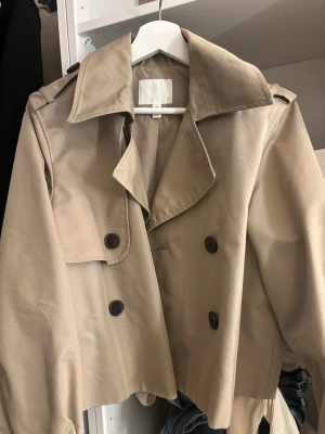 Kort beige trenchcoat med knappar från h&M - Snygg kort trenchcoat i beige med dubbelknäppning och bred krage. Klassisk design med axelklaffar och stora knappar framtill. Perfekt för dig som vill ha en stilren och tidlös jacka till vardagslooken. Är i ett bra skick, har en lite fläck som syns på bilden men man tänker inte på den på långt håll. Den är i en beige,grön färg 